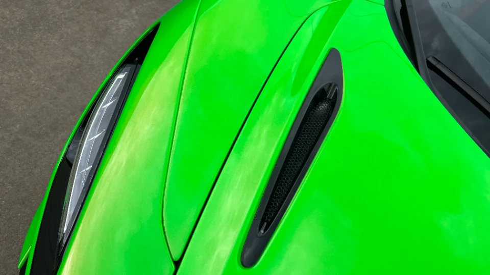 A close-up detail shot of a bright green supercar's front fender, featuring a sleek vent and part of the headlight.