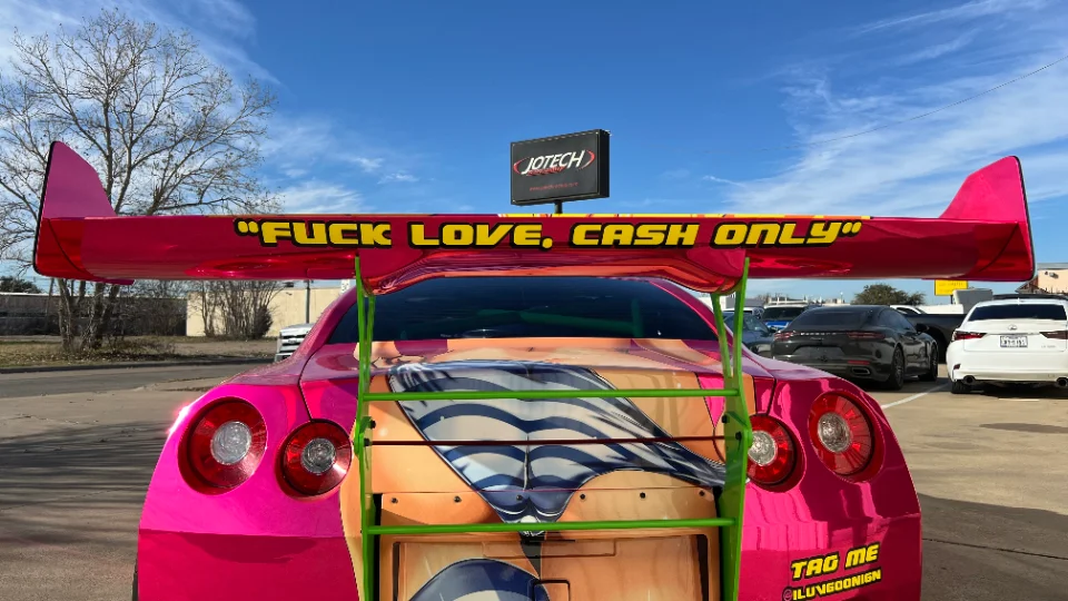 Rear view of a pink chrome wrapped Nissan GT-R with a large aftermarket wing and custom livery parked outdoors.