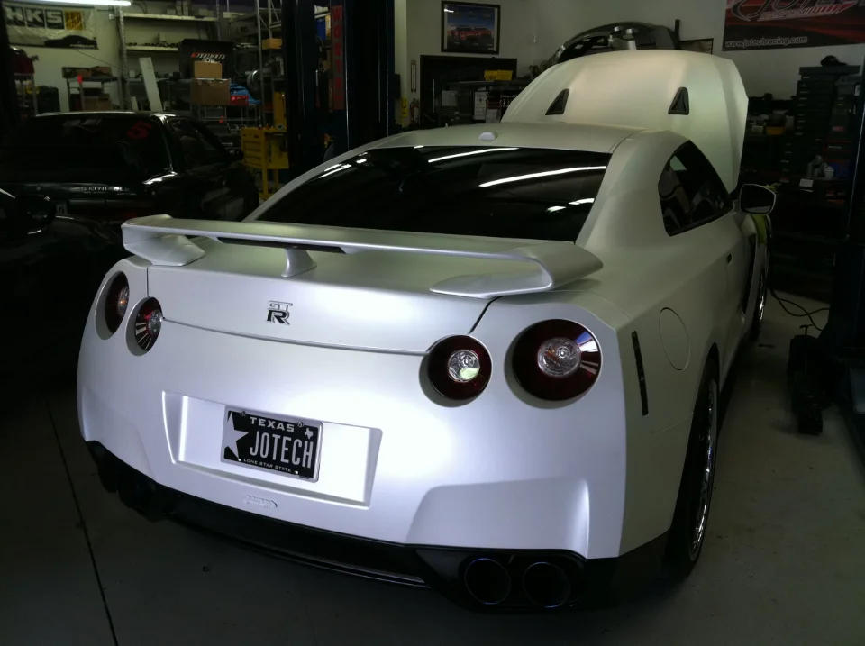 A white Nissan GT-R with its hood open, parked in a Jotech Motorsports shop with a custom Jotech license plate.