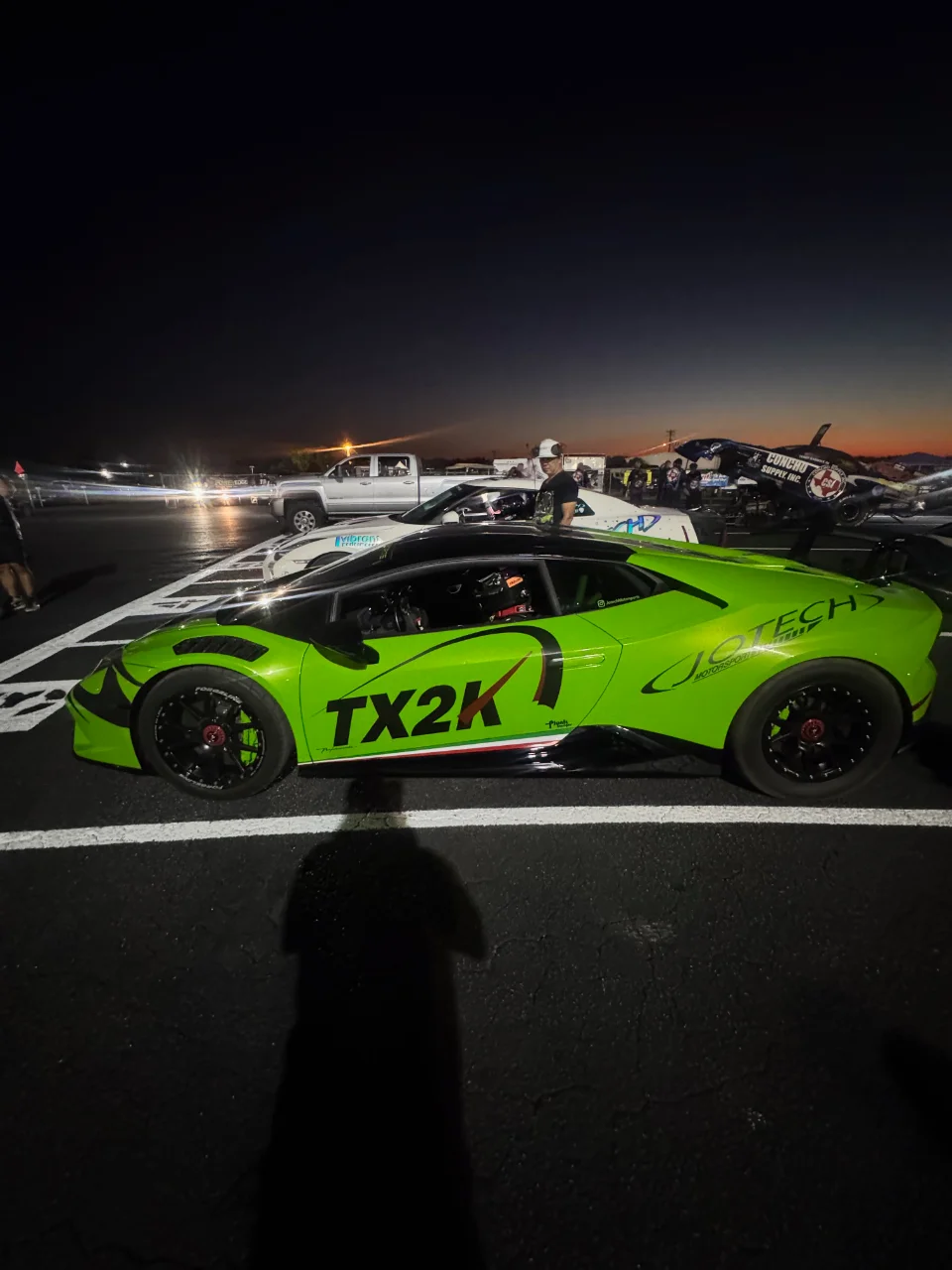 A bright green Lamborghini Huracan with TX2K livery parked at a drag strip at night, with other vehicles and people in the background.