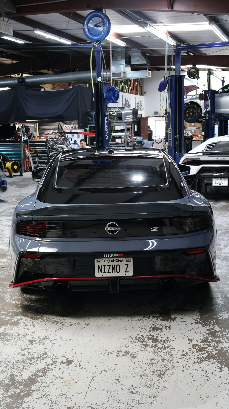 Rear view of a grey Nissan Z Nismo with a custom license plate 'NIZMO Z' parked in a performance tuning shop.