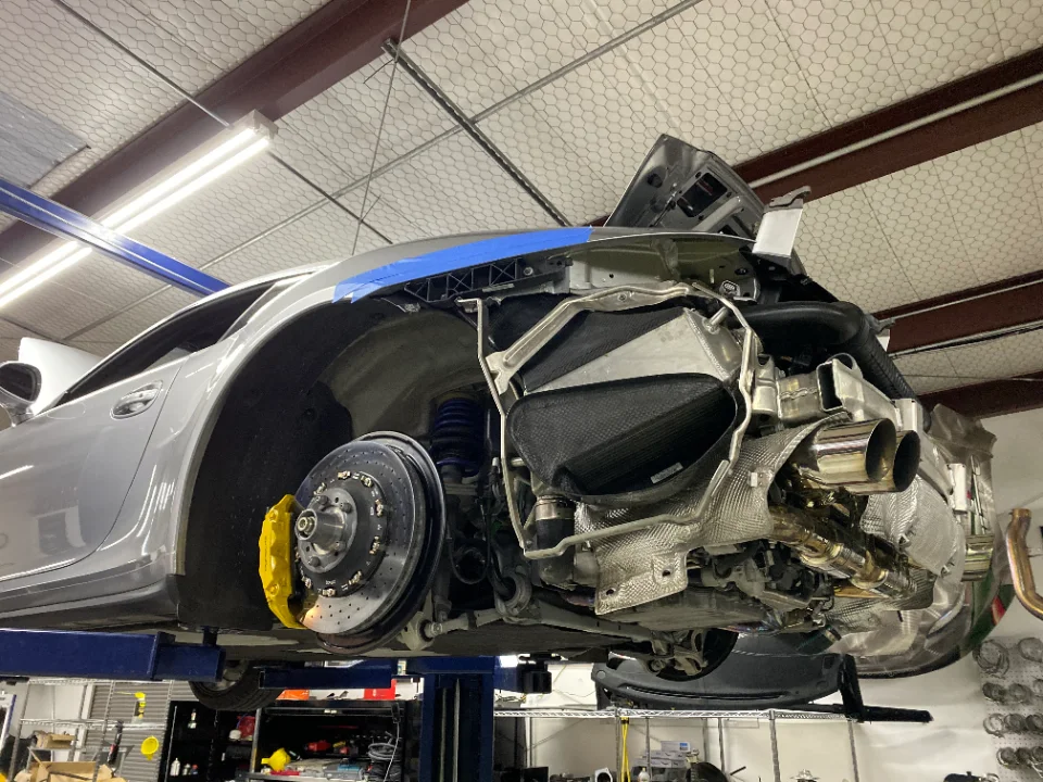 A silver Porsche 911 on a lift in a performance shop, with the rear bumper removed exposing the exhaust, turbo, and suspension.