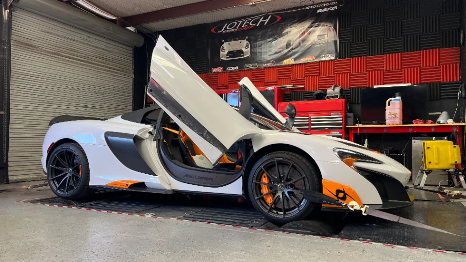 A white McLaren supercar with orange accents strapped to a dynamometer at Jotech Motorsports for performance testing.