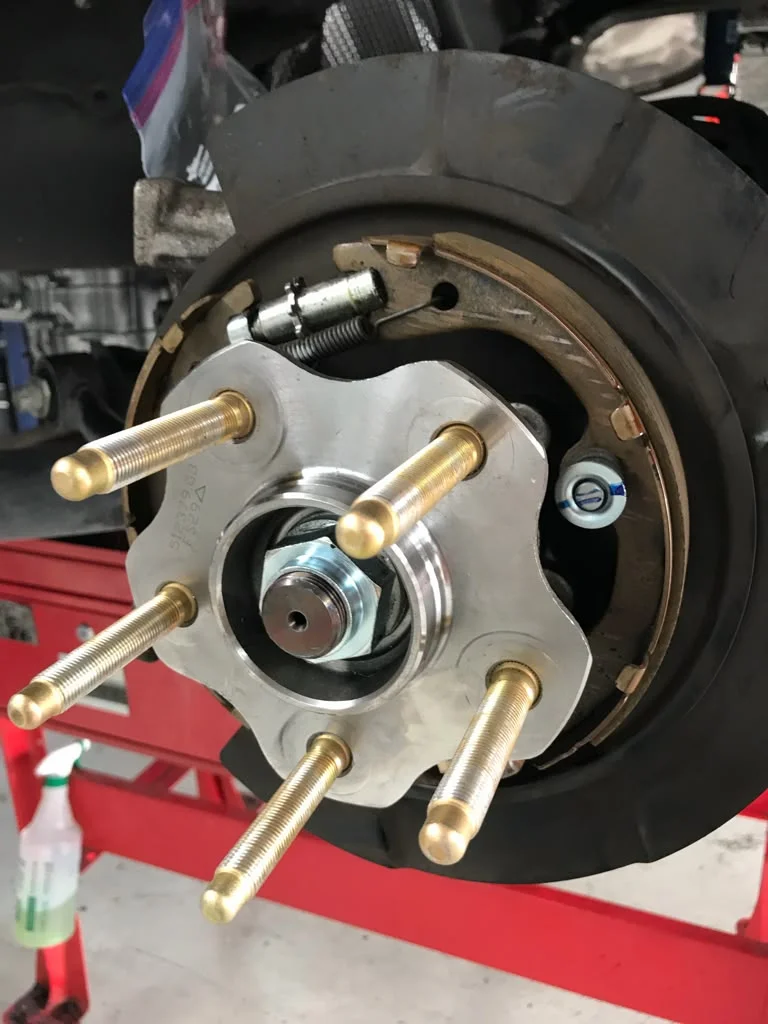 A close-up of a car's rear wheel hub and brake assembly with extended studs being installed in a shop environment.