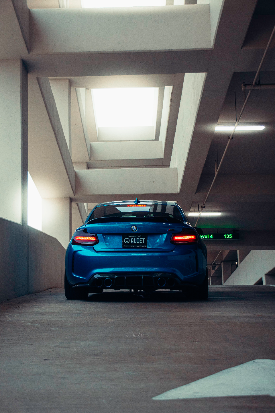 Rear view of a blue BMW M2 with quad exhaust and diffuser parked in a concrete parking garage.