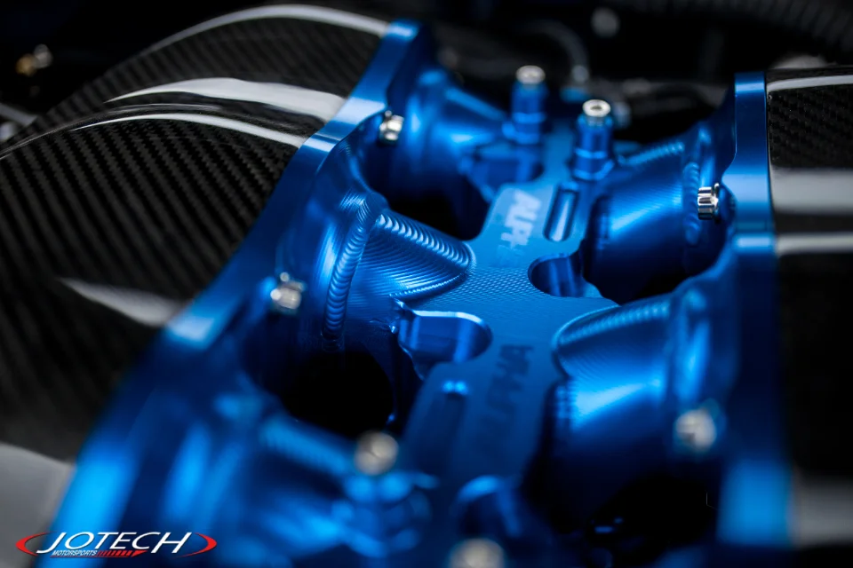 A close-up detail shot of a blue Alpha Performance intake manifold with carbon fiber components in an engine bay.