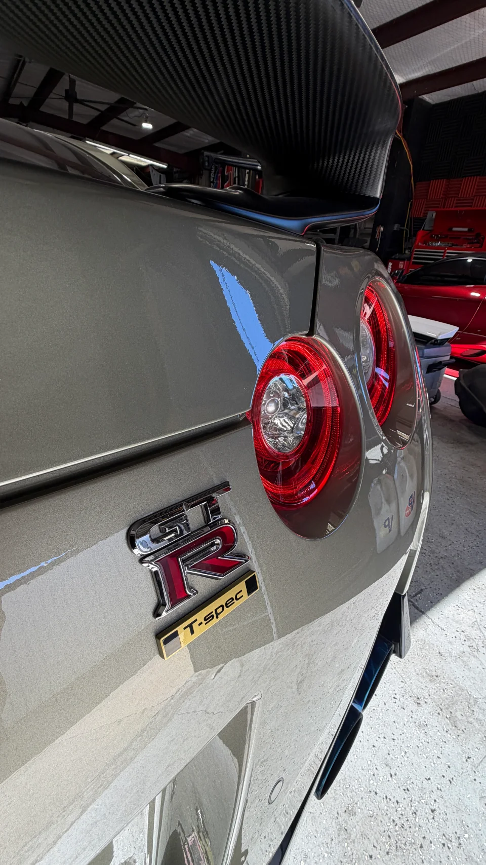 Rear view of a grey Nissan GT-R T-spec with a large carbon fiber wing and distinctive round taillights in a shop setting.