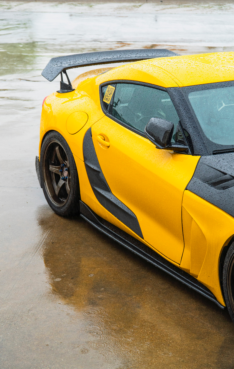 A vibrant yellow Toyota Supra with extensive carbon fiber aerodynamic modifications and aftermarket wheels, parked on a wet surface.