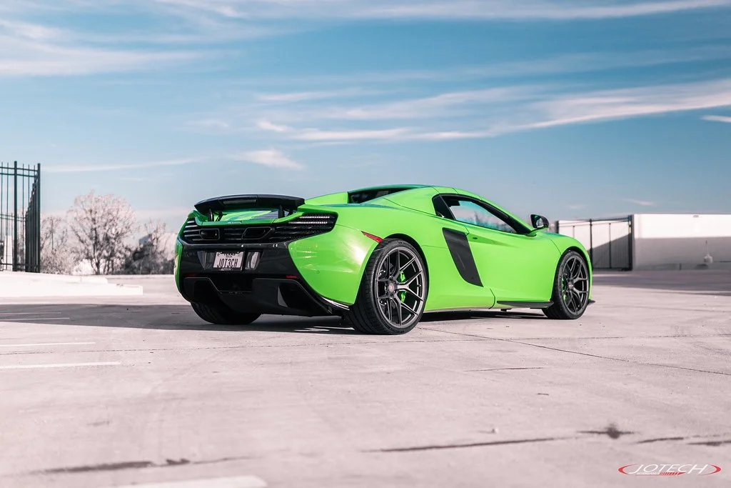 A vibrant green McLaren supercar with custom dark wheels and a rear spoiler, parked outdoors on a sunny day under a blue sky.