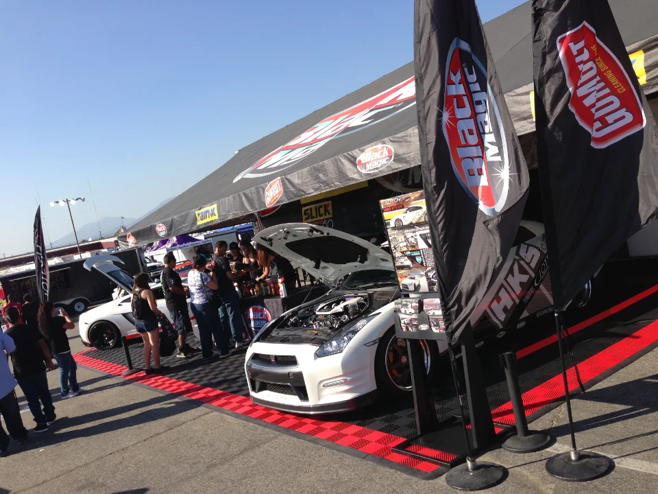 A white Nissan GT-R with its hood open on display at an outdoor car show event with vendor booths and a crowd.