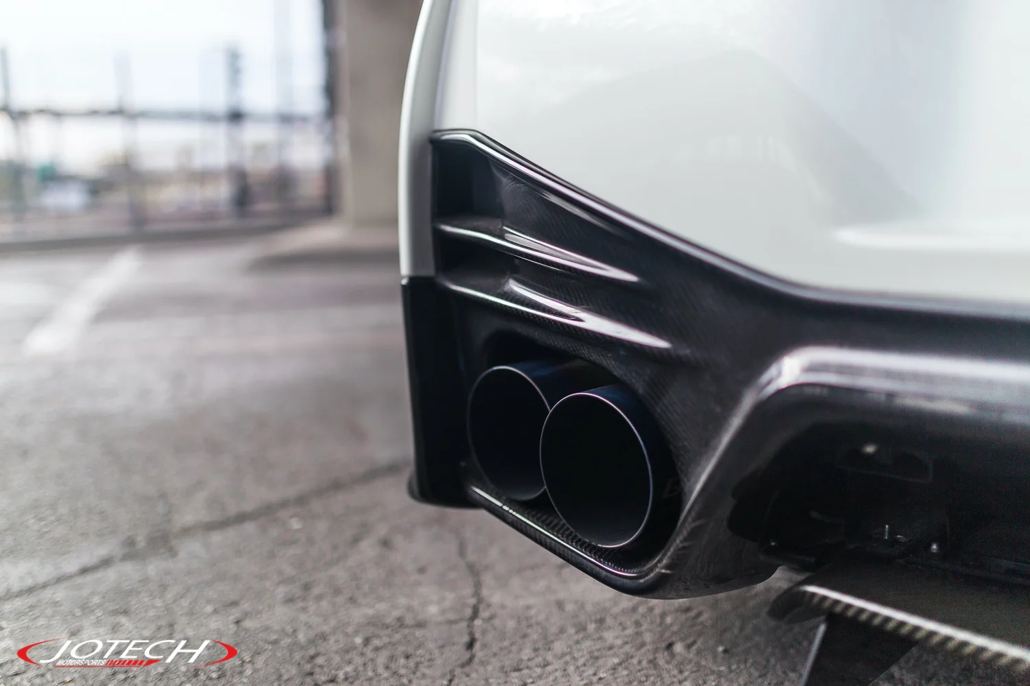 Close-up of a white car's rear bumper featuring dual exhaust tips and a carbon fiber diffuser, with Jotech branding.