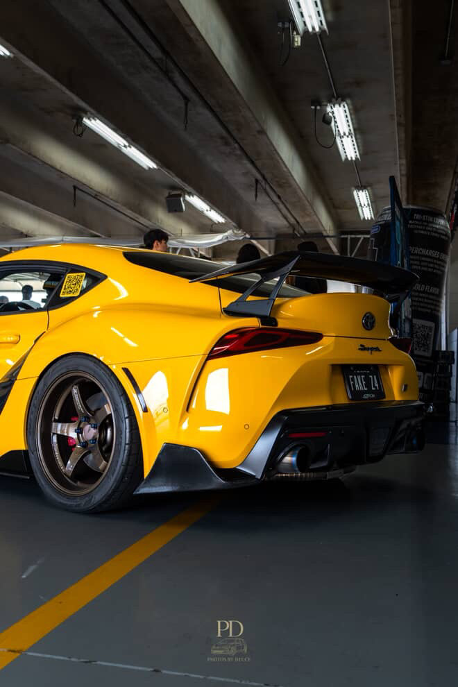 A yellow Toyota Supra A90 with aftermarket bronze wheels, a large carbon fiber wing, and a rear diffuser parked in a garage.