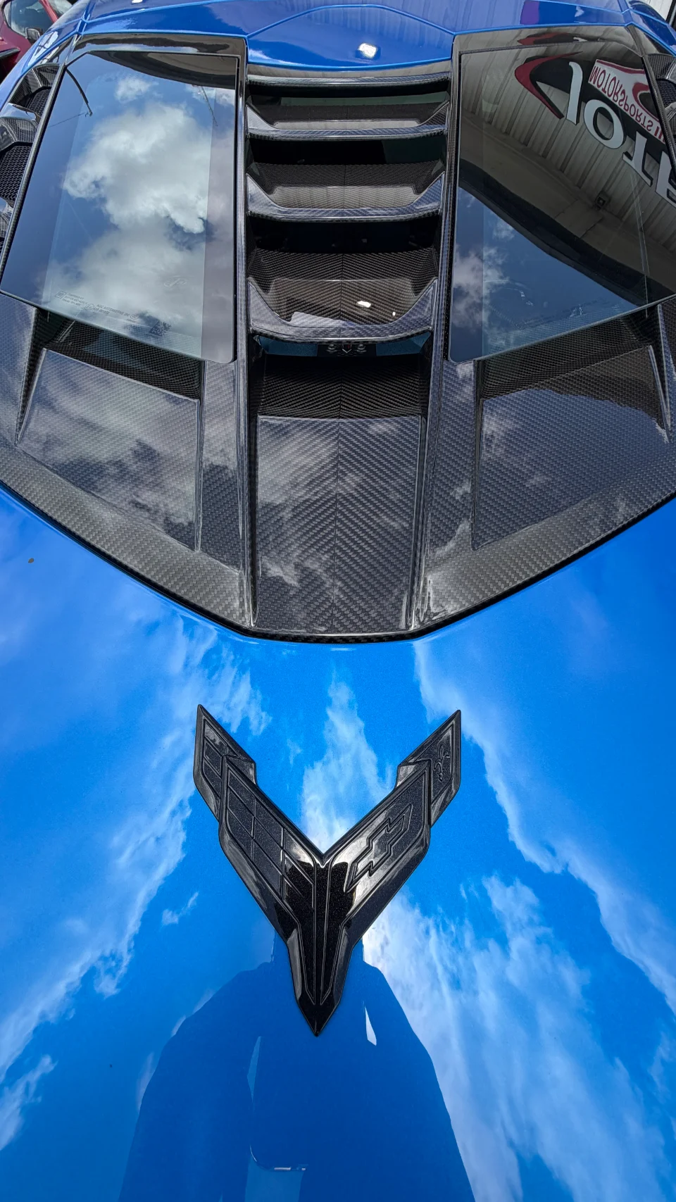 A close-up of the blue C8 Corvette's rear engine cover with carbon fiber accents and the black Corvette emblem, reflecting the sky.