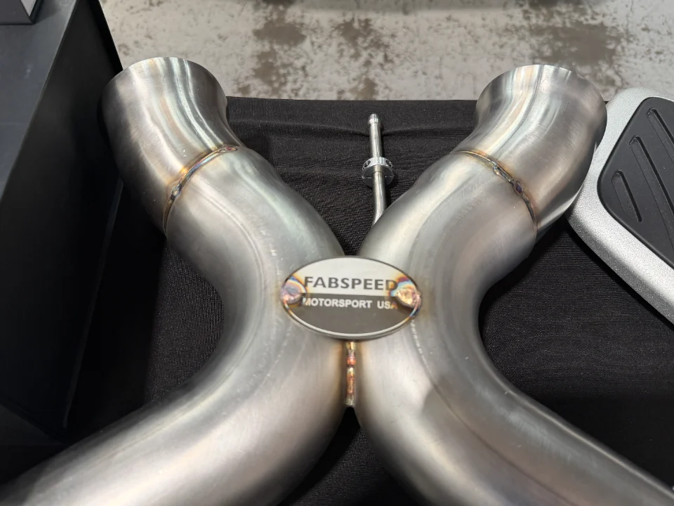 Close-up of a Fabspeed stainless steel X-pipe exhaust component with visible welds and branding.