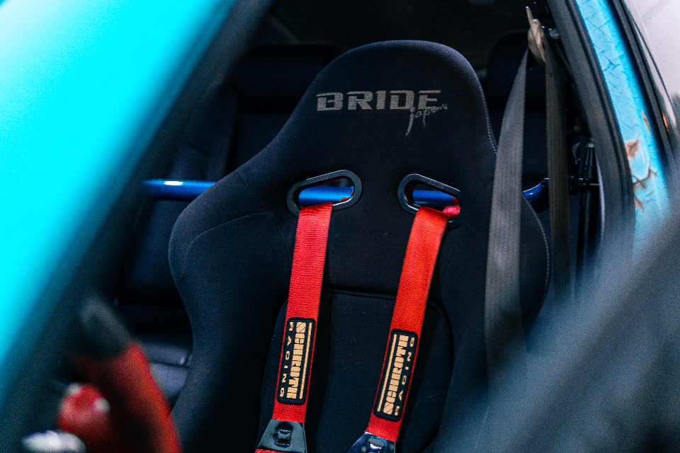 A close-up of a black Bride racing seat with red Schroth Racing harnesses and a blue roll bar in a car interior.