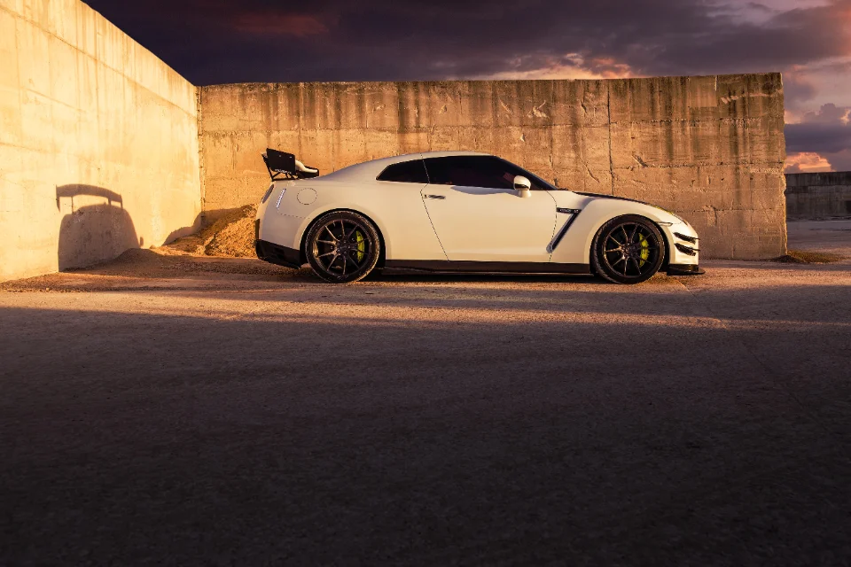 A white Nissan GT-R with aftermarket wheels and a large rear wing parked against a concrete wall at sunset.