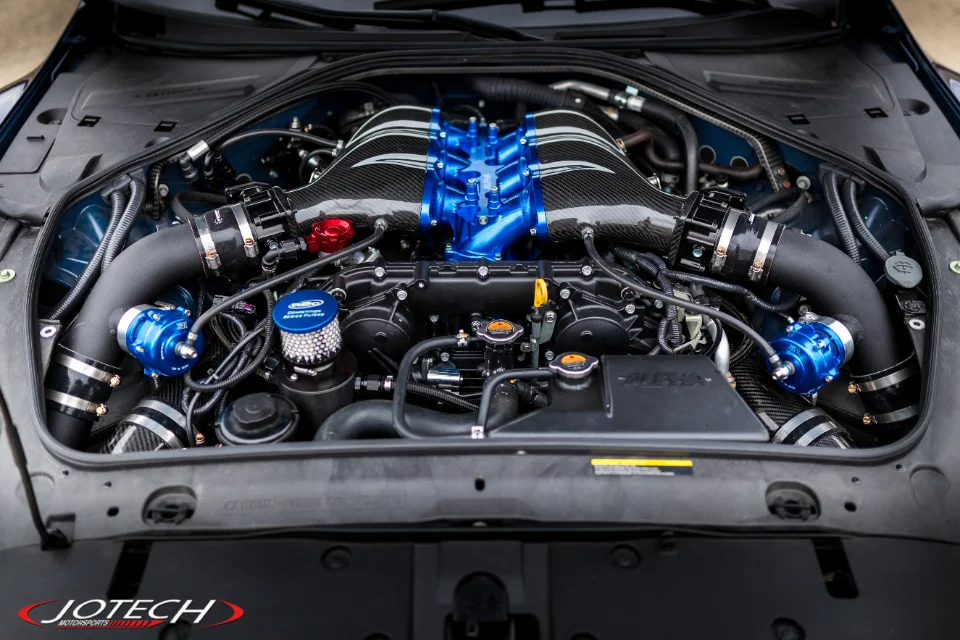 Close-up of a highly modified Nissan GT-R engine bay featuring a blue intake manifold, carbon fiber induction pipes, and blow-off valves by Jotech Motorsports.