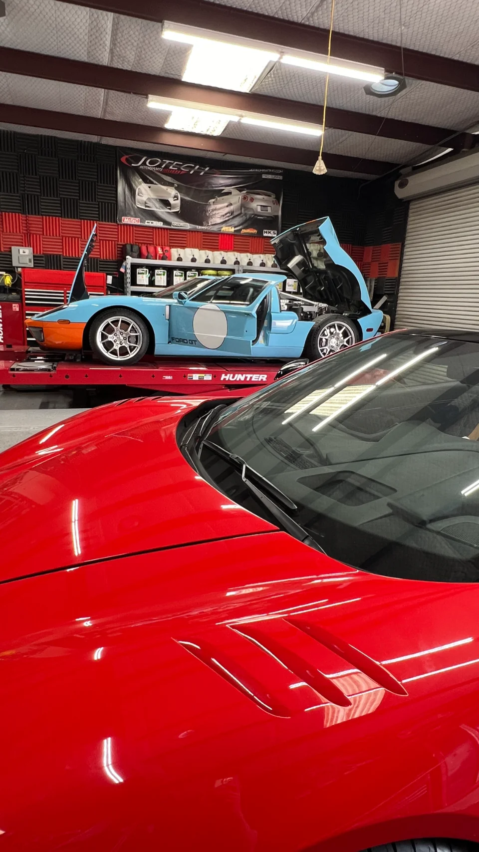 A light blue Ford GT with its engine bay open on a red car lift in a Jotech Motorsports shop, with a red Corvette in the foreground.