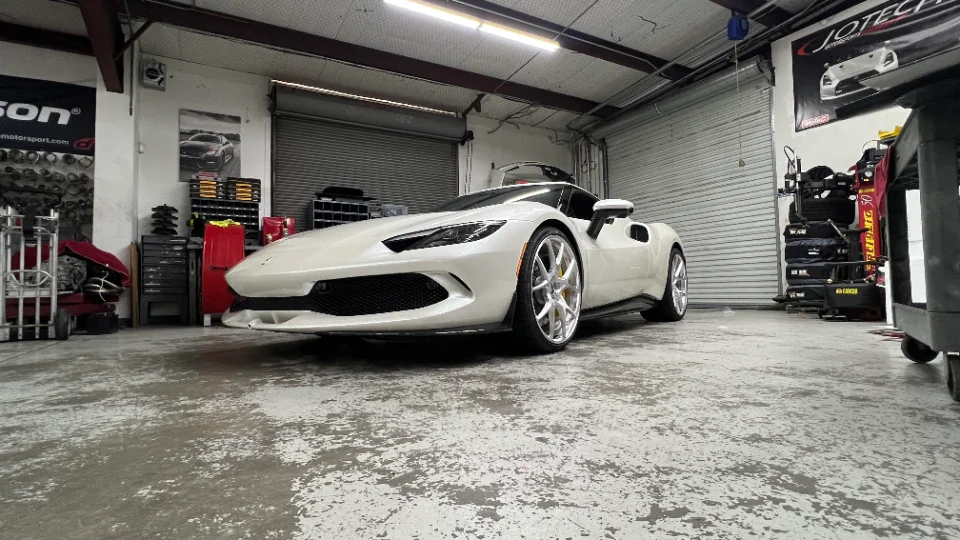 A white Ferrari 296 GTB sports car with custom silver wheels parked inside a Jotech Motorsports performance tuning shop.