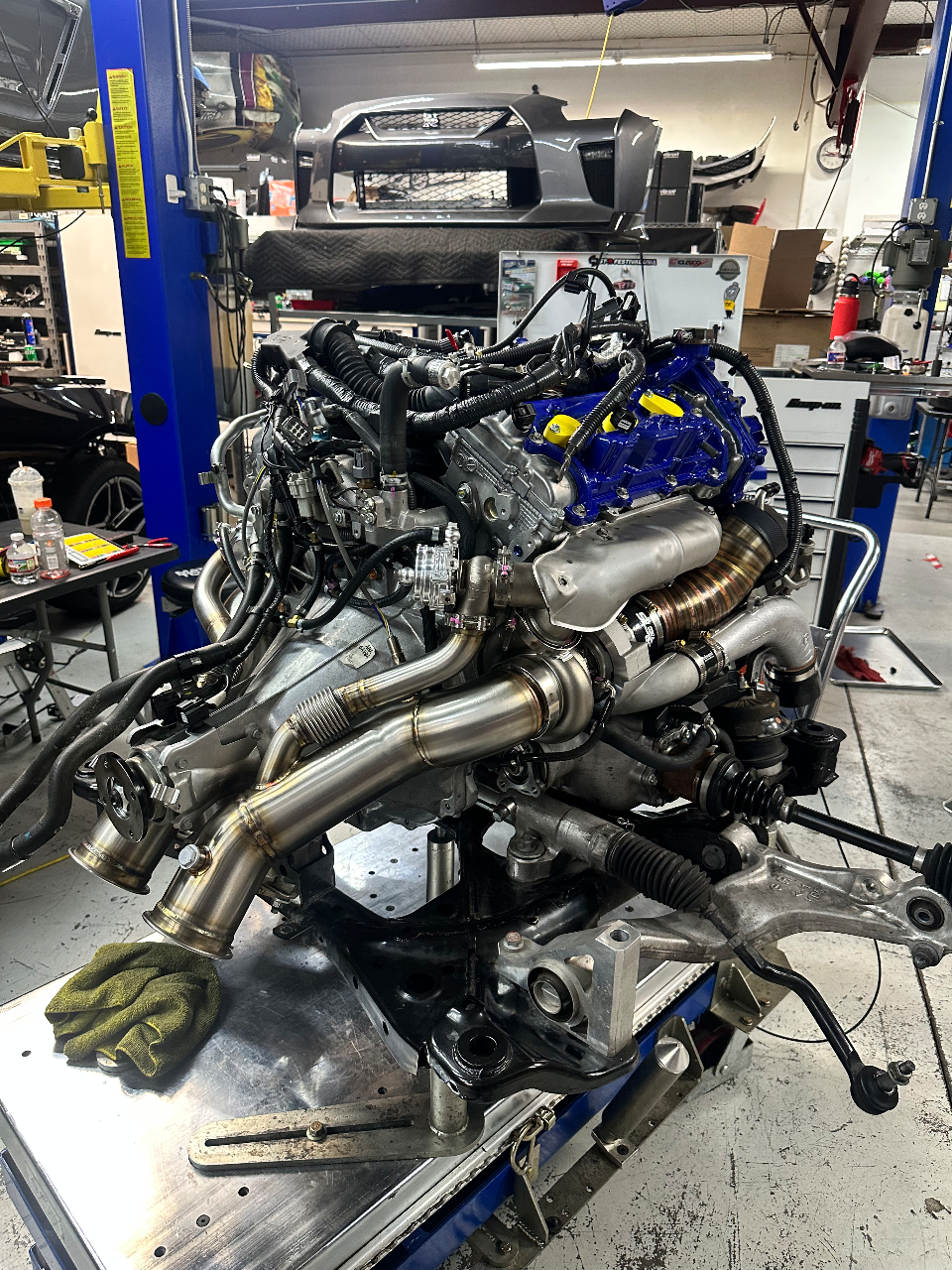 A high-performance V6 engine with custom blue valve covers, turbochargers, and exhaust manifolds on a stand in a Jotech Motorsports shop.