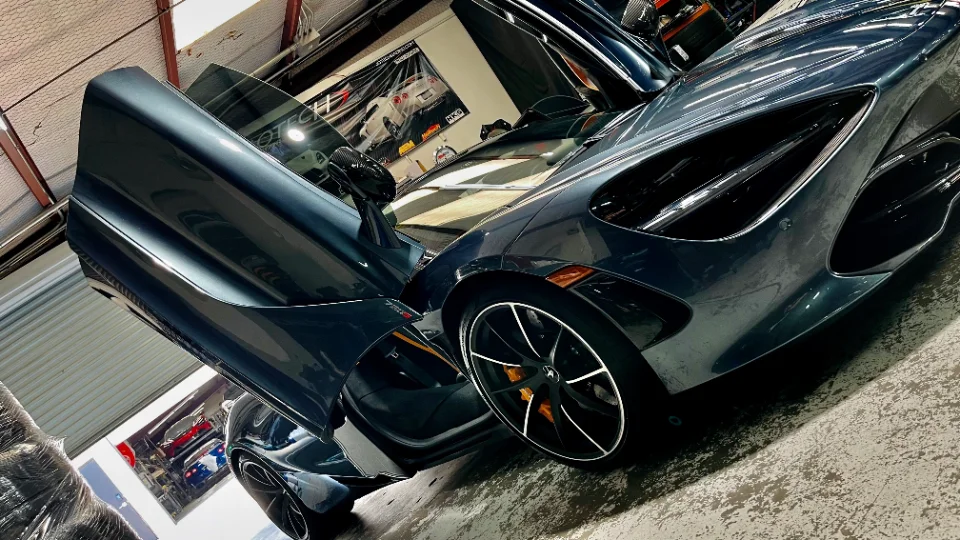 A dark grey McLaren 720S with its distinctive butterfly doors open, parked inside a Jotech Motorsports performance tuning shop.