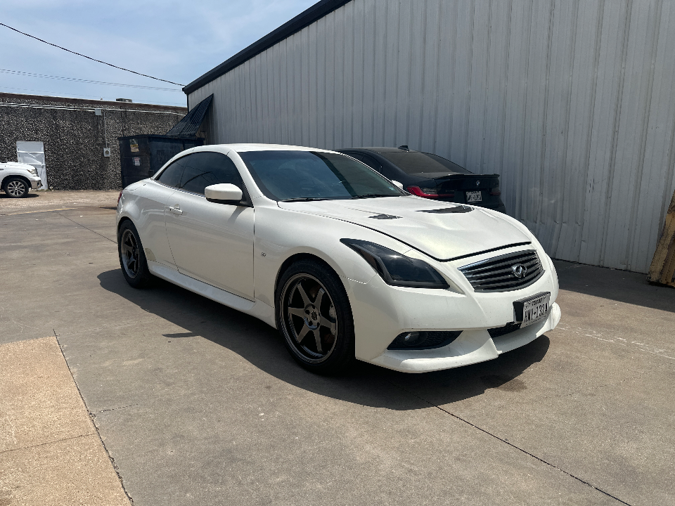 White Infiniti G37 coupe with aftermarket wheels and a custom hood parked outside an industrial building on a sunny day.