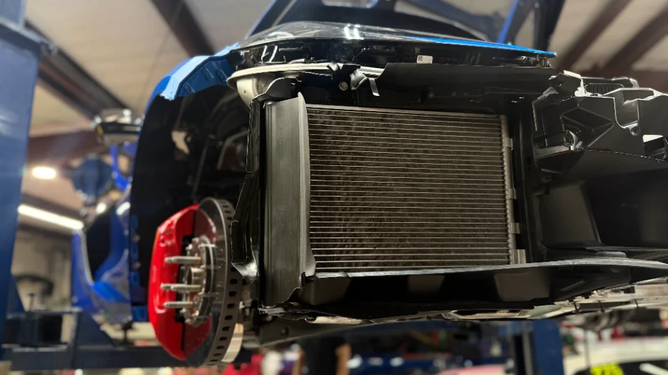 A blue C8 Corvette on a lift in a shop, with the front bumper removed, revealing the radiator and red brake calipers.
