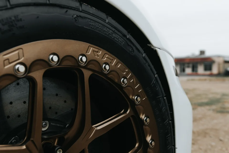 Close-up of a bronze multi-spoke racing wheel with a performance tire and drilled brake rotor on a white car.