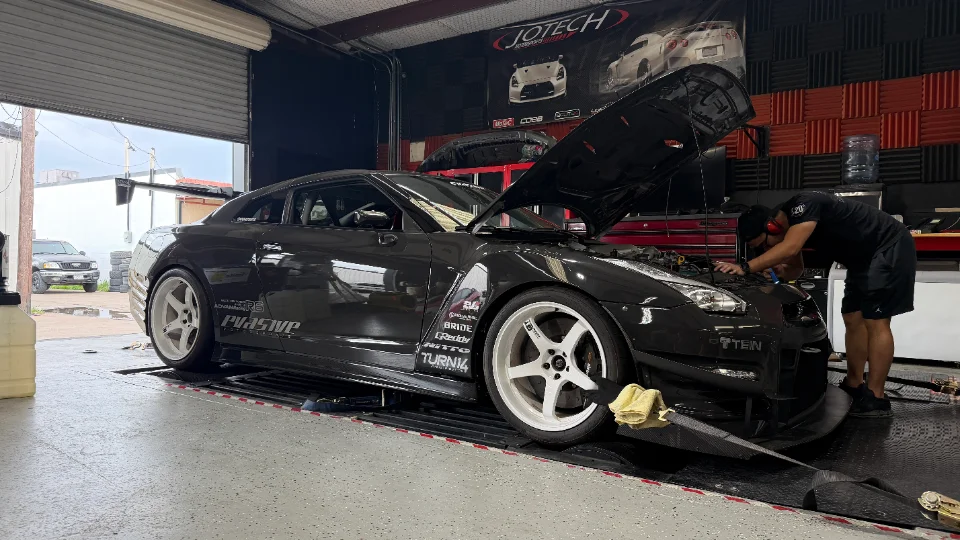 A black Nissan GT-R R35 on a dyno at Jotech Motorsports with a mechanic working on the engine bay.