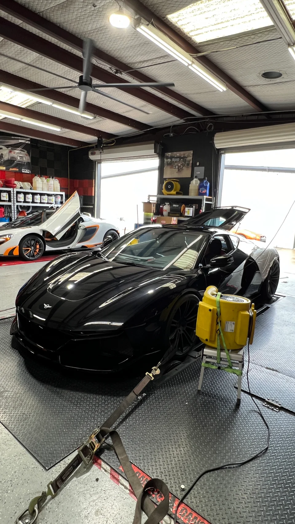 Black McLaren supercar strapped to a dynamometer in a performance tuning shop, with another McLaren in the background.