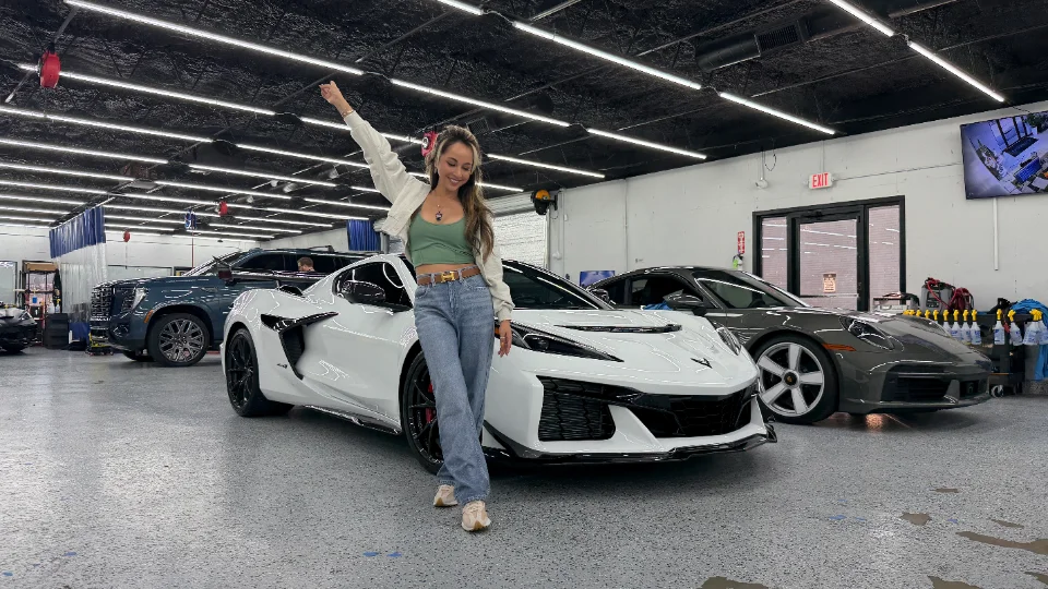 A woman poses in a clean car shop with a white Chevrolet Corvette C8 Z06 and a dark grey Porsche 911 in the background.
