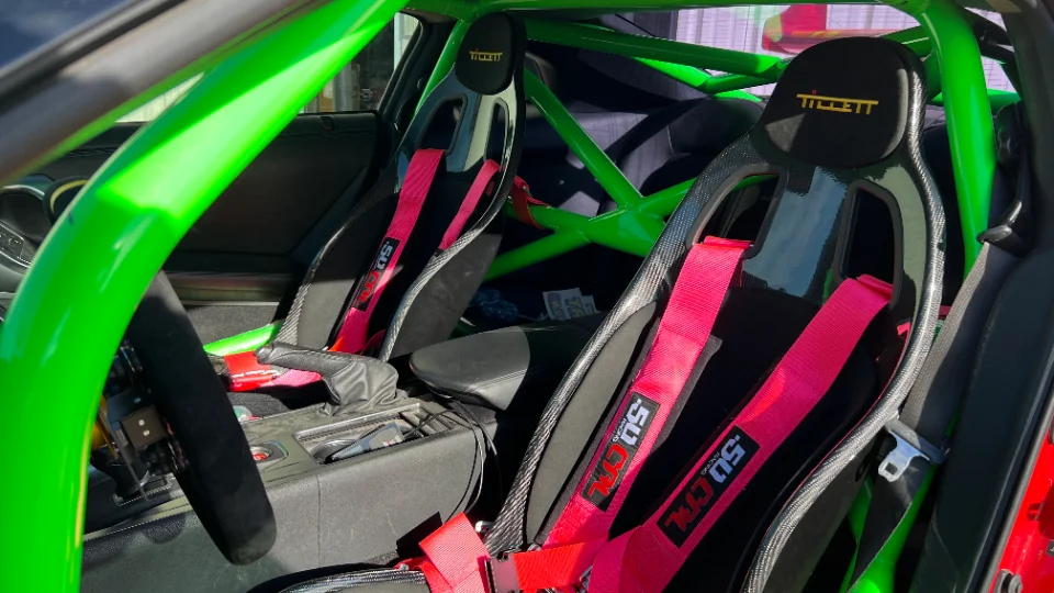 A close-up of a car's interior featuring a bright green roll cage, Tillett racing seats, and red 50 GTL harnesses.