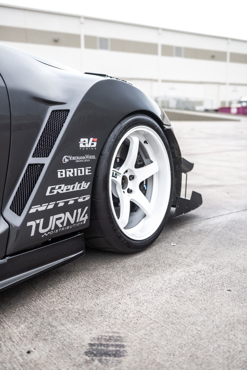 Close-up of a grey widebody car's front wheel and fender, featuring white multi-spoke wheels, performance tires, carbon fiber side skirt, and sponsor decals.