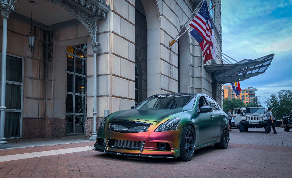 A custom iridescent wrapped Infiniti G37 sedan with a front splitter and aftermarket wheels parked in front of a building.