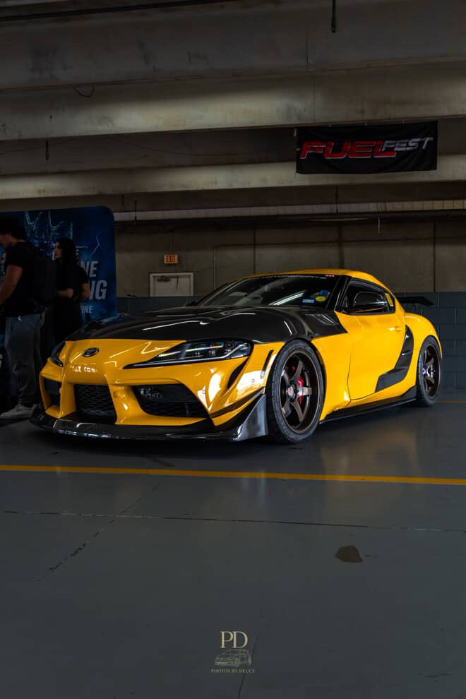 A highly modified yellow and black Toyota Supra with carbon fiber aero parts and aftermarket wheels at an indoor event.