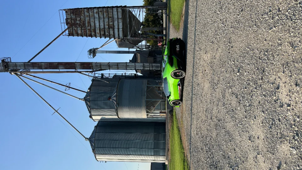 A vibrant green Lamborghini supercar parked on a gravel road next to large grain silos under a clear blue sky.