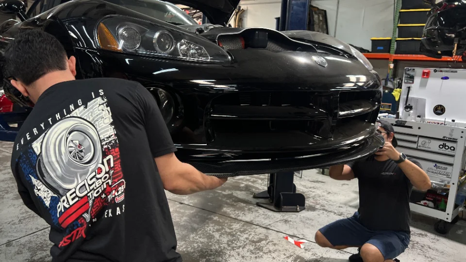 Two mechanics installing a carbon fiber front splitter on a black Dodge Viper in a performance tuning shop.