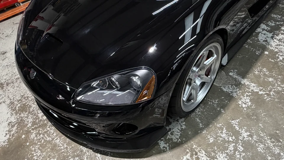 Close-up of a shiny black Dodge Viper's front end, featuring a carbon fiber splitter and aftermarket wheels on a concrete floor.