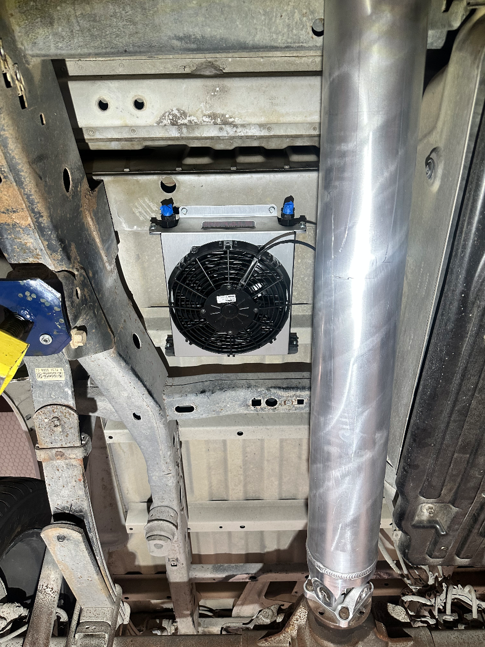 Underside view of a vehicle showing a newly installed transmission cooler with an electric fan and a custom driveshaft.