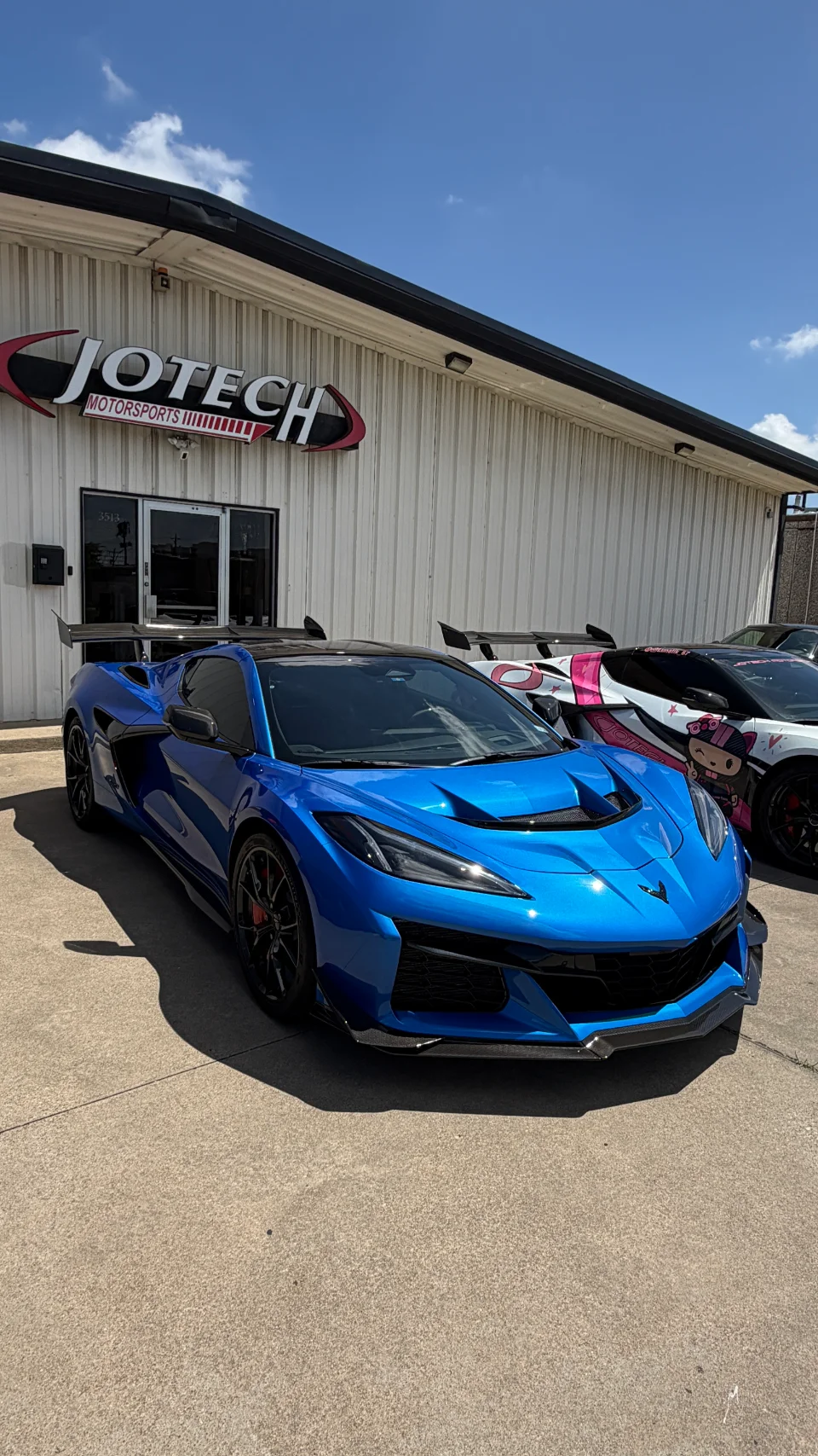 A blue Chevrolet Corvette C8 Z06 with a large rear wing parked outside Jotech Motorsports, with another custom car behind it.