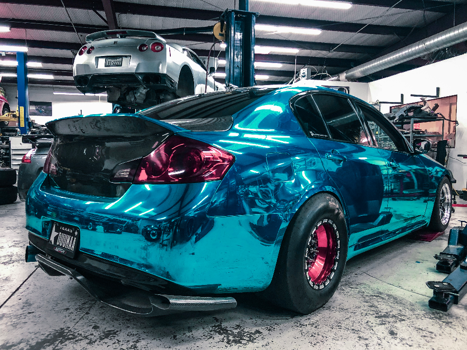 A blue chrome wrapped Infiniti G37 sedan with drag slicks and a carbon fiber spoiler in a performance shop, with a silver Nissan GT-R on a lift.