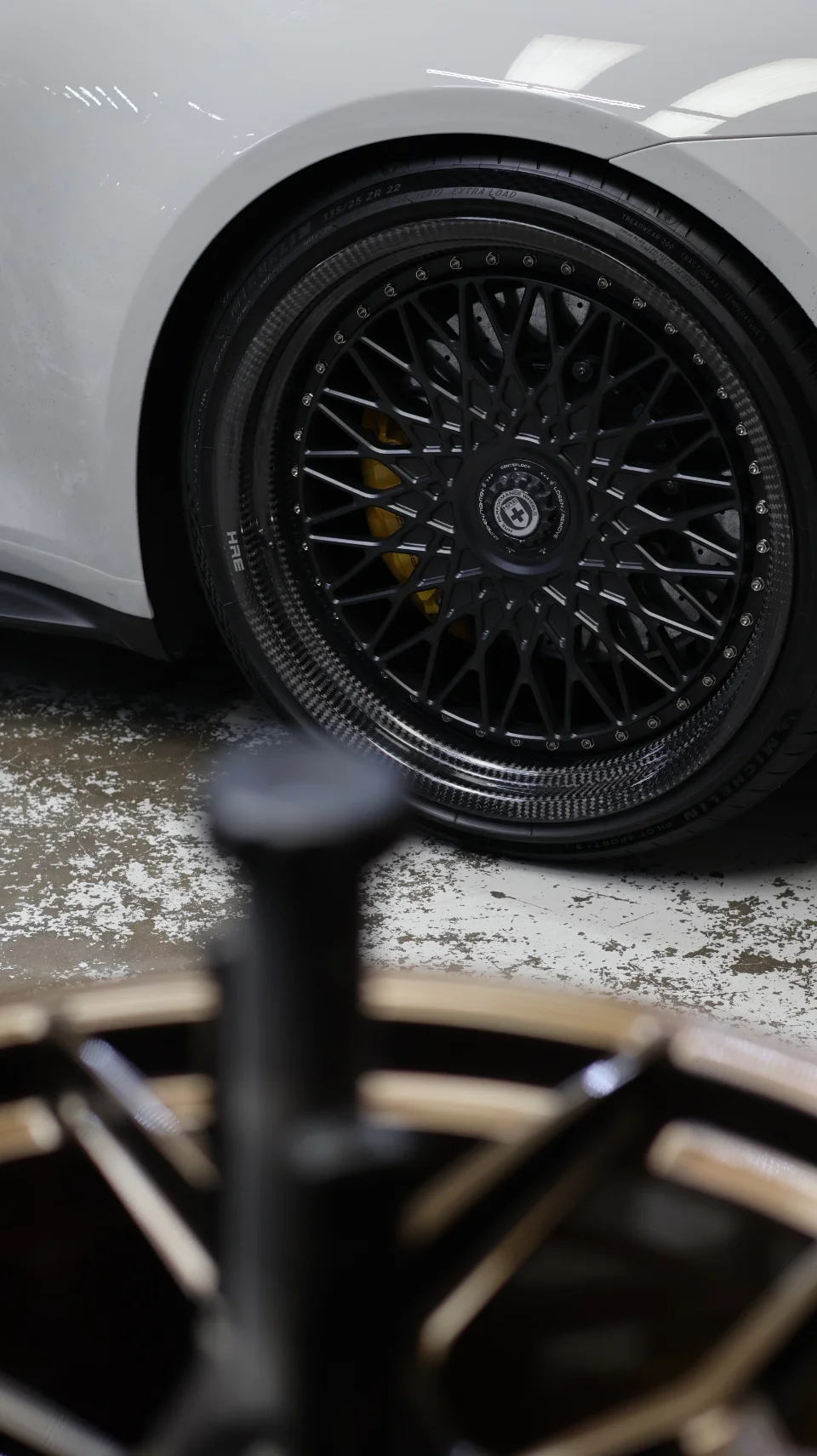 Close-up of a white car's front wheel, featuring black HRE performance wheels with carbon fiber barrels and yellow brake calipers.