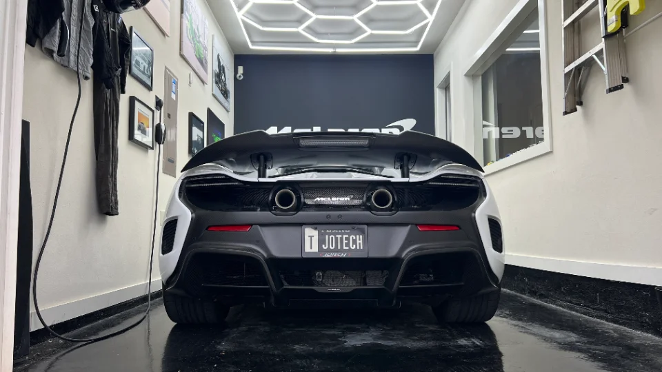 Rear view of a black and white McLaren supercar with a carbon fiber wing and diffuser, parked in a well-lit Jotech Motorsports shop.