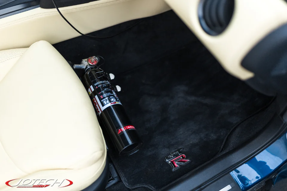 A black fire extinguisher rests on a black Nissan GT-R branded floor mat in the passenger footwell of a car with light-colored leather seats.