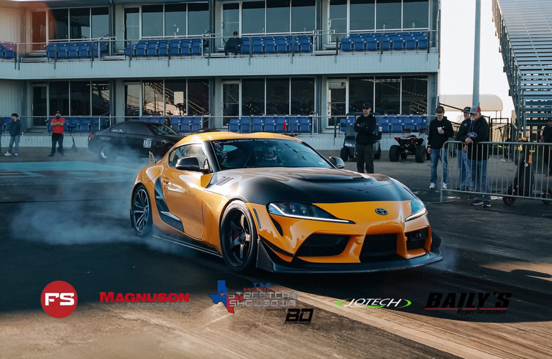 A yellow Toyota Supra with a widebody kit performs a burnout on a drag strip, with grandstands and spectators in the background.