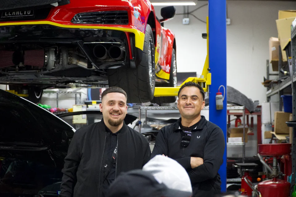 Two mechanics stand in a car shop with a red Corvette C8 on a lift, showcasing its twin-turbo setup and exhaust.