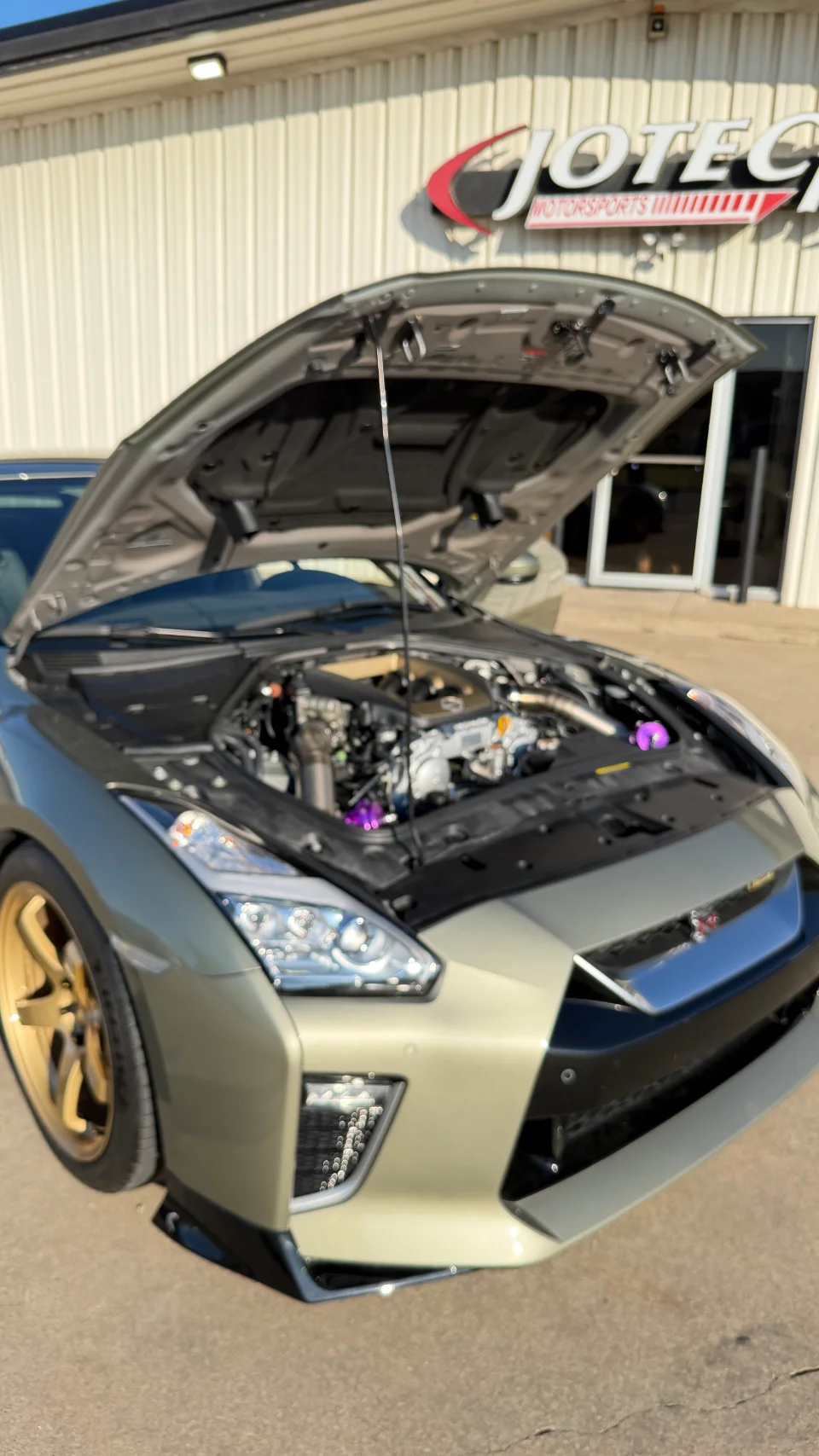 A Nissan GT-R with its hood open, showcasing the engine bay, parked in front of the Jotech Motorsports shop.