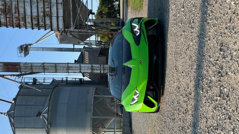 A vibrant green Lamborghini Huracan parked on a gravel road, set against a backdrop of large industrial silos under a clear blue sky.