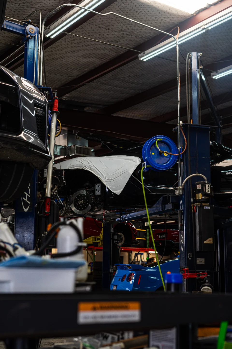 Multiple performance cars on lifts in a busy automotive shop, with a blue Nissan GT-R visible in the background.