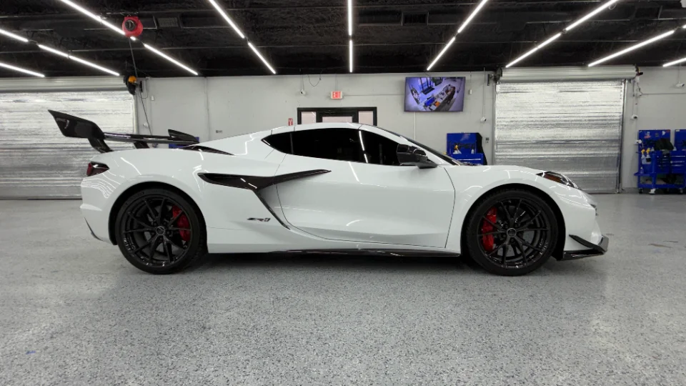White Chevrolet Corvette C8 Z06 with a large carbon fiber rear wing and red brake calipers parked in a clean shop.
